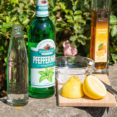 Das Bild zeigt die Zutaten für den Pfeffi Twist: Soda, Orangenlikör, eine Pfeffi Flasche, Eis und Zitrone