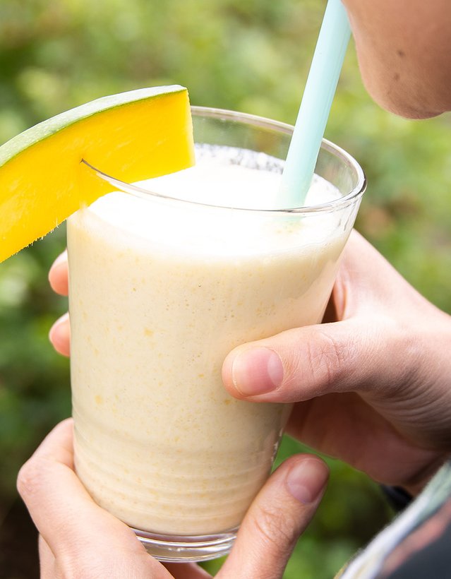 Das Bild zeigt eine Person, die an einem Glas mit milchiger Flüssigkeit und einer Mangoscheibe am Rand aus einem Strohhalm trinkt