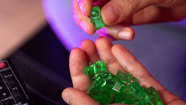 Man sieht eine Hand mit grünen Gummibärchen darin. Im Hintergrund ist eine Fernbedienung zu sehen
