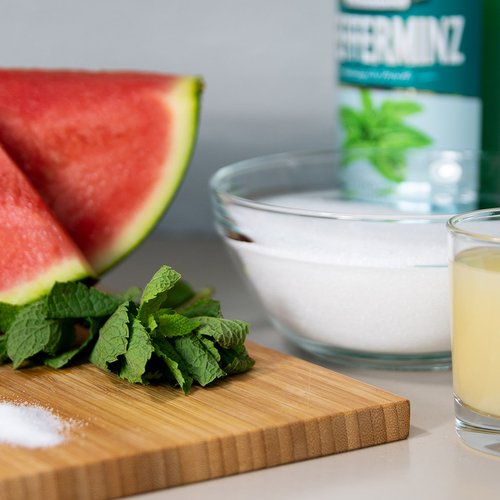 Das Bild zeigt eine Wassermelone, eine Flasche Pfeffi, Minze, Zitronensaft und Zucker auf einem Schneidebrett.