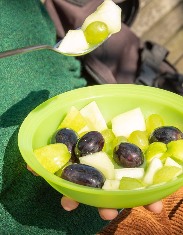 Das Bild zeigt eine Person, die draußen im Grünen aus einem Plastikteller einen Obstsalt isst
