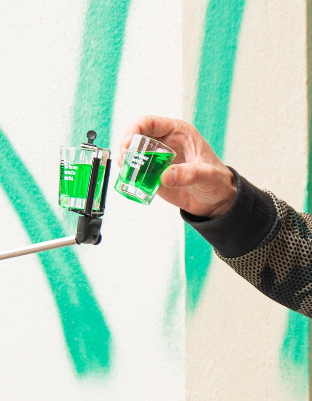 Das Bild zeigt, wie ein Pinnchen an einem Selfiestick zu einem zweiten Pinnchen geführt wird, das eine Person in der Hand hält
