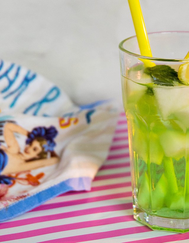 Das Bild zeigt einen grünen Cocktail mit einer Zitrone und einem gelben Strohhalm auf einer weiß-pink gestreiften Tischdecke