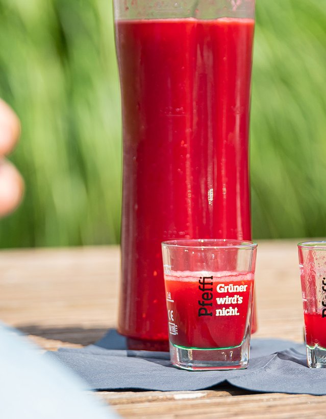 Man sieht eine mit roter Flüssigkeit gefüllte Flasche, davor eine Person, die auf einer Bank sitzt mit einem Pinnchen in der Hand. Daneben liegt eine Sonnenbrille