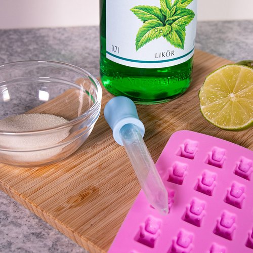 Das Bild zeigt eine Silikonform für Gummibärchen, eine Zitrone, Gelierzucker und eine Flasche Pfeffi