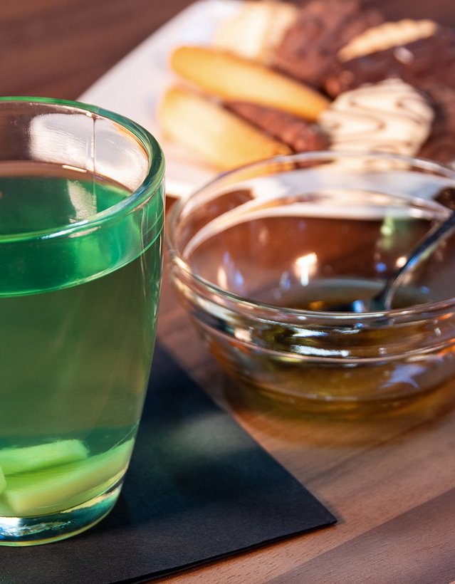 Pfeffi Rezept Pfeffiminztee Das Bild zeigt einen Teetasse mit grünem Inhalt, einer Zitronenscheibe und einer Schale Kekse im Hintergrund