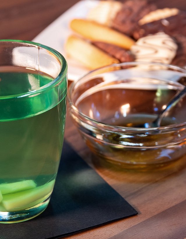 Das Bild zeigt einen Teetasse mit grünem Inhalt, einer Zitronenscheibe und einer Schale Kekse im Hintergrund