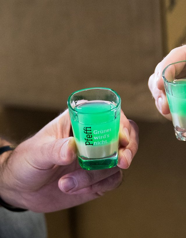 Pfeffi Rezept Doppeldecker Das Bild zeigt zweoi menschen die mit einem Pinnchen anstoßen, die Flüssigkeit darin ist geschichtet: unten ist die milchig, oben grün