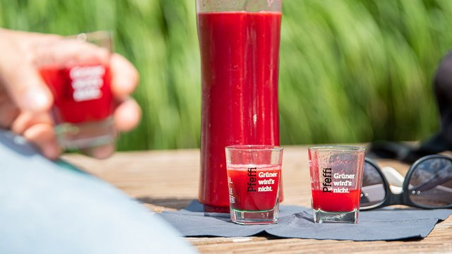 Man sieht eine mit roter Flüssigkeit gefüllte Flasche, davor eine Person, die auf einer Bank sitzt mit einem Pinnchen in der Hand. Daneben liegt eine Sonnenbrille