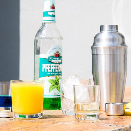 Das Bild zeigt einen Cocktailshaker, eine Flasche Pfeffi, Orangensaft, Blue Curacao und Eiswürfel