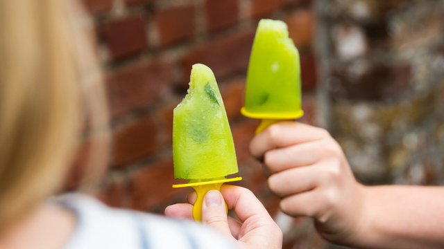 Das Bild zeigt eine Hand, die ein grün-gelbes Eis am Stil vor sich hält