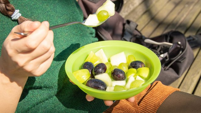 Das Bild zeigt eine Person, die draußen im Grünen aus einem Plastikteller einen Obstsalt isst