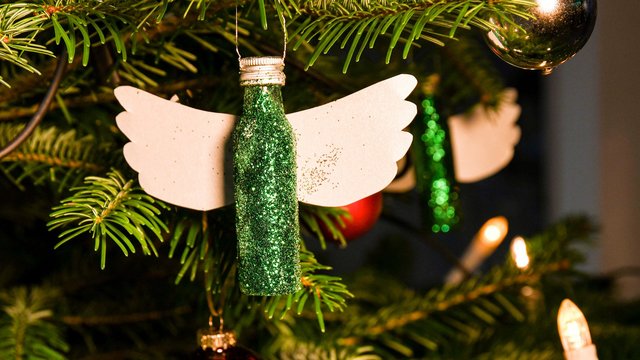 Das Bild zeigt eine mit grünem Glitzer bedeckte Pfeffi Mini Flasche, die mit zwei Flügeln an der Rückseite versehen wurde, sodass sie wie eine Engelsfigur aussieht. Sie hängt an einem Christbaum.