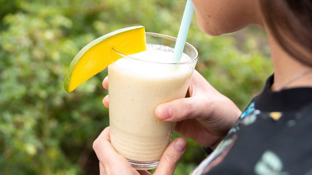 Das Bild zeigt eine Person, die an einem Glas mit milchiger Flüssigkeit und einer Mangoscheibe am Rand aus einem Strohhalm trinkt