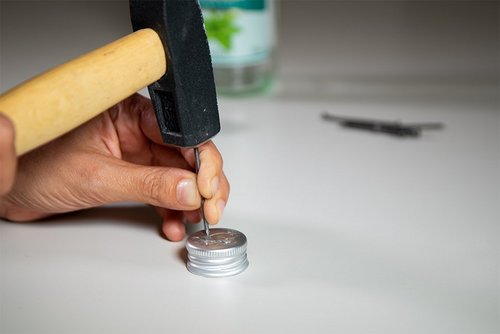 Das Bild zeigt, wie in einen Pfeffi Flaschendeckel mittels Hammer und Nagel ein Loch geschlagen wird
