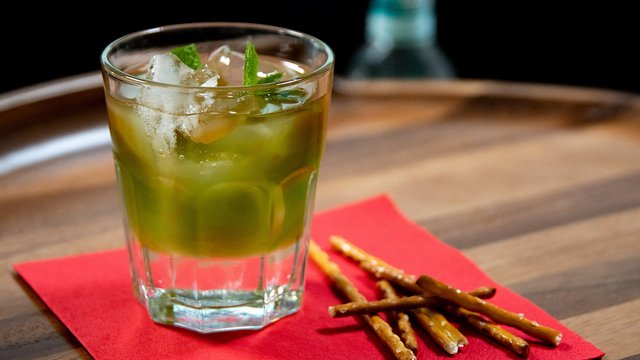 Das Bild zeigt einen grünen Pfeffi Cocktail auf einer roten Serviette nebst Salzstangen