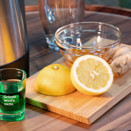 Pfeffi Rezept Pfeffiminztee Zutaten Man sieht ein Stück Ingwerwurzel, eine aufgeschnittene Zitrone, eine Schale mit Honig und ein Pfeffi Pinnchen neben einem Wasserkocher im Anschnitt