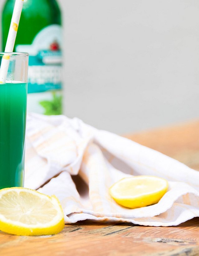 Pfeffi Rezept Grünling Das Bild zeigt einen grünen Cocktail mit einer Flasche Pfeffi im Anschnitt auf einem Holzitsch