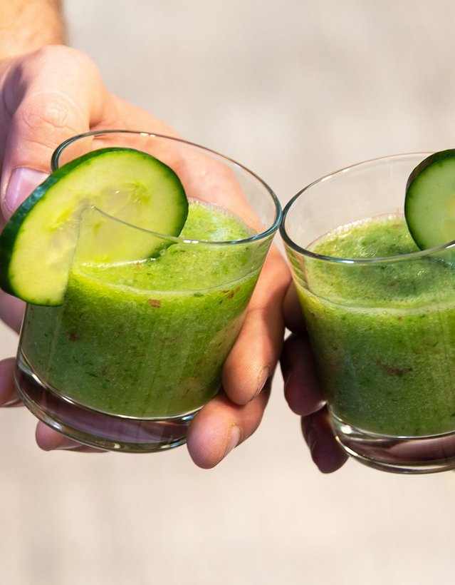 Pfeffi Rezept Minti Das Bild zeigt die Hände zweier Menschen, die mit je einem Glas mit grüner Flüssigkeit und Gurkenscheibe am Rand anstoßen