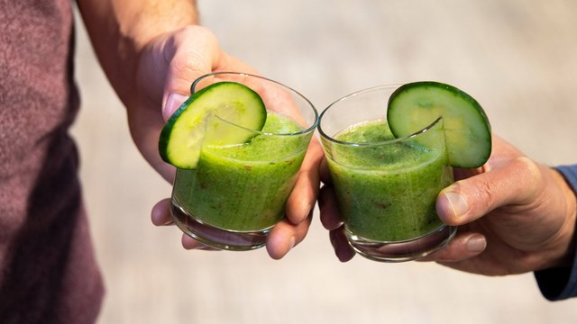 Das Bild zeigt die Hände zweier Menschen, die mit je einem Glas mit grüner Flüssigkeit und Gurkenscheibe am Rand anstoßen 