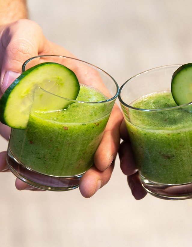 Pfeffi Rezept Minti Das Bild zeigt die Hände zweier Menschen, die mit je einem Glas mit grüner Flüssigkeit und Gurkenscheibe am Rand anstoßen