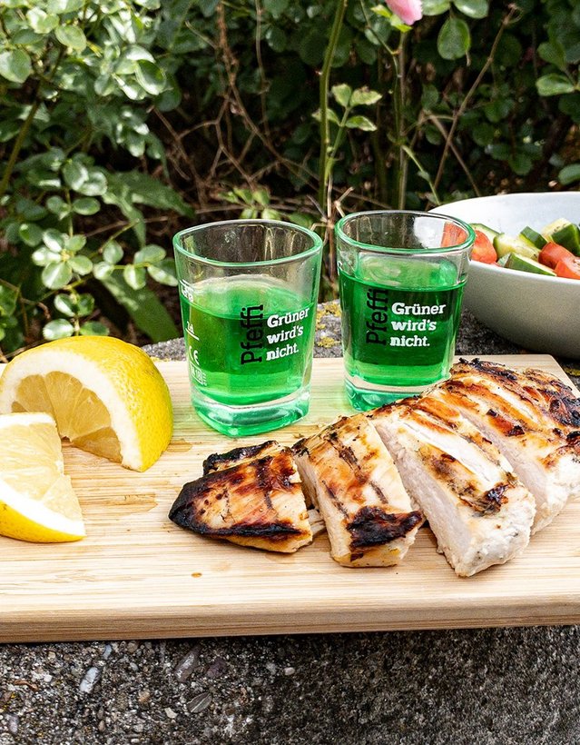 Pfeffi Rezept Pfeffis heiße Chicks Das Bild zeigt eine gegrillte Hühnerbrust die aufgeschnitten auf einem Brettchen liegt neben einer Schüssel Salat, ein paar Pfeffi Pinnchen und einer Zitrone