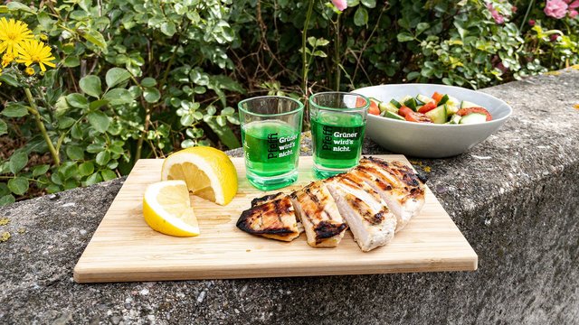 Das Bild zeigt eine gegrillte Hühnerbrust die aufgeschnitten auf einem Brettchen liegt neben einer Schüssel Salat, ein paar Pfeffi Pinnchen und einer Zitrone