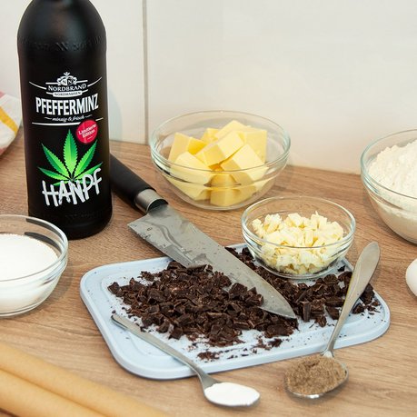 Pfeffi Rezept Hanpf Cookies Zutaten Das Bild zeigt eine Flasche Pfeffi Hanpf, sowie diverse weitere Backzutaten vor einem Schneidebrett mit gehackter Schokolade