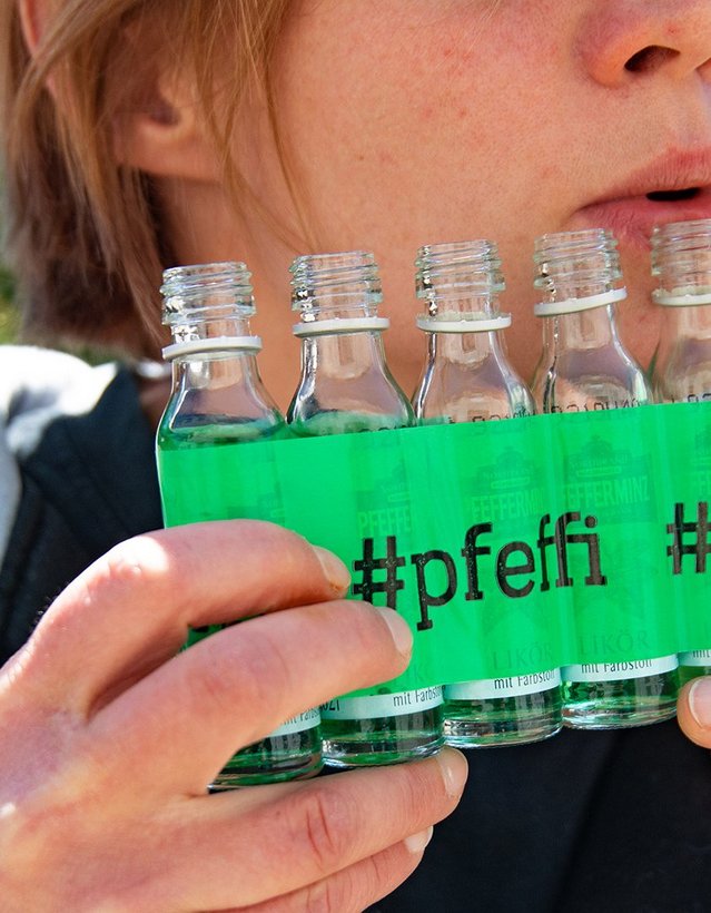 DIY: Pfeffi Panflöte Das Bild zeigt mehrere Pfeffi Mini Flaschen, die mit Klebeband in einer Reihe zusammengeklebt sind. Eine Person scheint darauf, wie auf einer Panflöte zu spielen.
