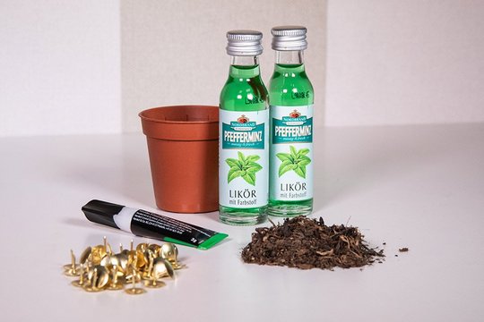 Das Bild zeigt Klebstoff, zwei Pfeffi Mini Flaschen, ein paar Reißzwecken, einen Blumentopf und ein wenig Erde