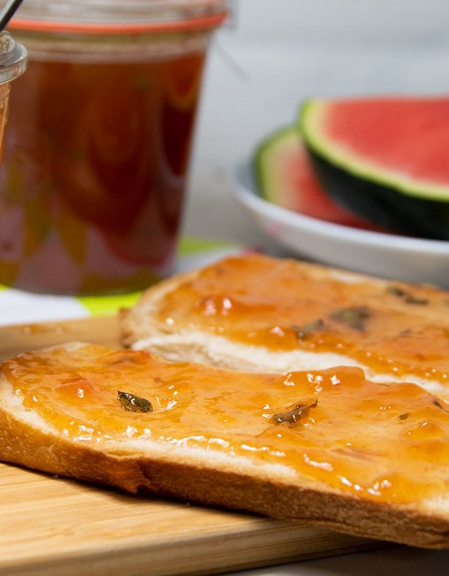 Das Bild zeigt ein Marmeladenglas nebst eines mit Marmelade beschmierten Toasts, im Hintergrund liegt eine Wassermelone.