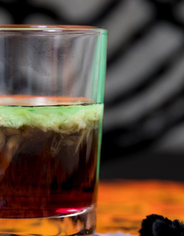 Das Bild zeigt einen dunklen Cocktail mit grünem "Topping"