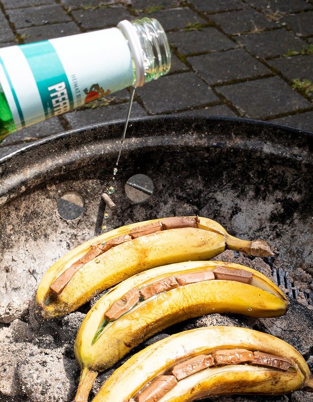 Das Bild zeigt einen Grill, in dessen Glut angeschnittene Bananen liegen, die mit Schokolade gefüllt sind, während jemand Pfeffi aus der Flasche über die Banane gießt 