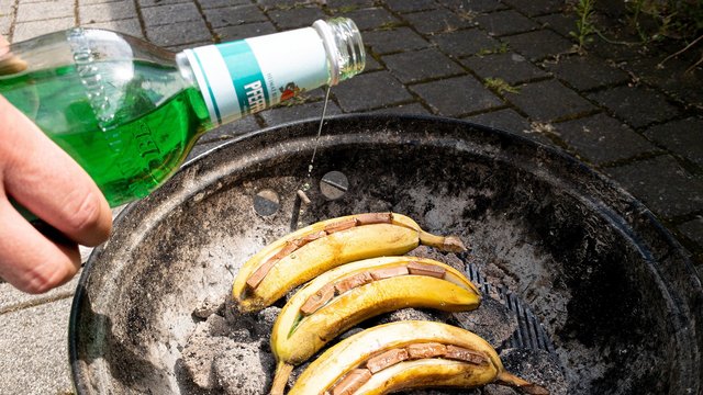 Das Bild zeigt einen Grill, in dessen Glut angeschnittene Bananen liegen, die mit Schokolade gefüllt sind, während jemand Pfeffi aus der Flasche über die Banane gießt 