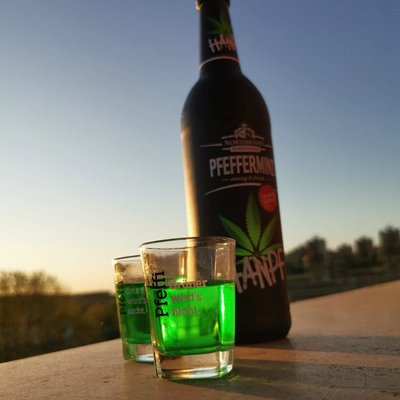 Hanpf Fanbild 1 Das Bild zeigt eine Hanpf Flasche nebst zwei gefüllten Pinnchen draußen auf einem Holztisch