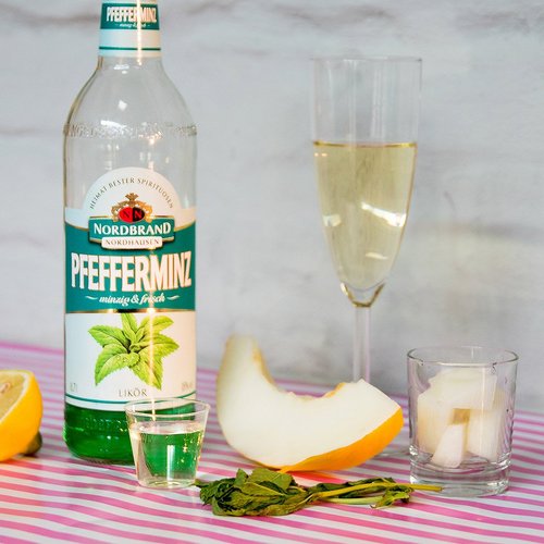 Pfeffi Rezept Flotter Dreier Zutaten Das Bild zeigt eine Pfeffi Flasche, ein Glas Sekt und ein Stück Melone, sowie eine Zitrone im Anschnitt