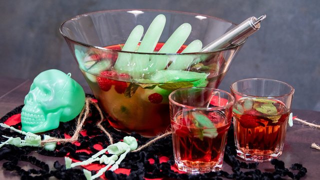 Das Bild zeigt eine Bowle, die von Halloweendeko umgeben ist. Aus der Bowle ragt eine Hand aus grünem Eis